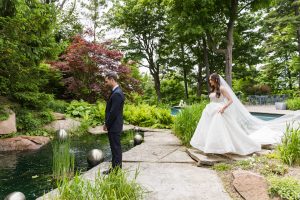 First look setup: bride approaching groom from behind in lush garden beside pond