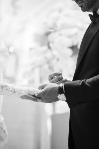 Black-and-white close-up of groom about to place wedding band onto bride’s finger during the ceremony, highlighting the ring and lace sleeve