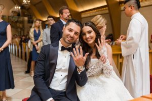 Newlywed couple in church pews smiling and holding up their wedding rings toward the camera, guests and priest softly blurred in the background