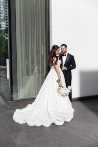 Bride and groom standing side-by-side in formal attire on a modern city sidewalk, bride holding a bouquet as they pose against a sleek glass-and-stone building