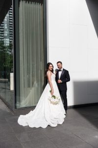 Full-length exterior portrait of bride and groom in elegant wedding attire standing beside urban architecture, their long train displayed on pavement