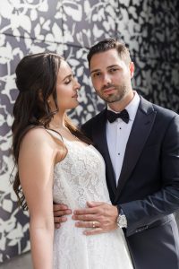Close-up portrait of bride gazing at groom while he looks ahead, both posed in front of abstract patterned wall in sunlight