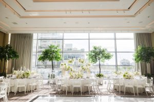 Wide shot of sunlit wedding reception venue with round tables, white chairs, lush floral centerpieces, and tall indoor trees by floor-to-ceiling windows