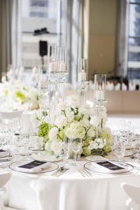 Elegant floral centerpiece featuring white roses, hydrangeas, greenery, and tall glass candles illuminating a reception table
