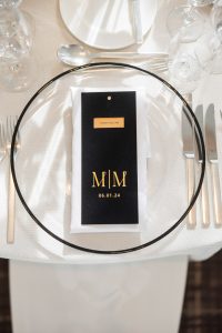 Overhead shot of a single place setting with personalized black-and-gold menu labeled “M | M 06.01.24” resting atop crisp white napkin and clear charger plate