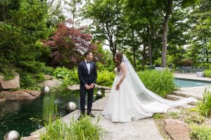 Bride and groom smiling at each other by pond, bride’s veil and gown in full view