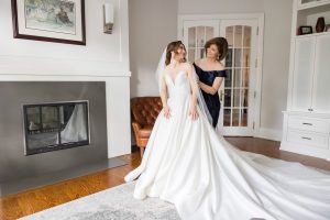 Bride smiles at her mom while getting dressed in a white gown at home.