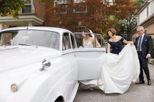 Bride enters a white vintage car with help from her parents.