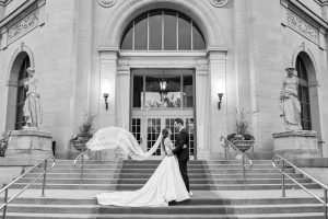 Bride’s veil blows dramatically as the couple embraces on Liberty Grand’s staircase.
