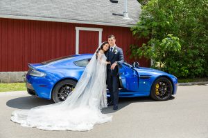Bride and groom pose beside a blue Aston Martin in front of a red barn