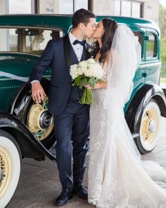 Bride and groom kiss in front of a vintage green car holding a white bouquet